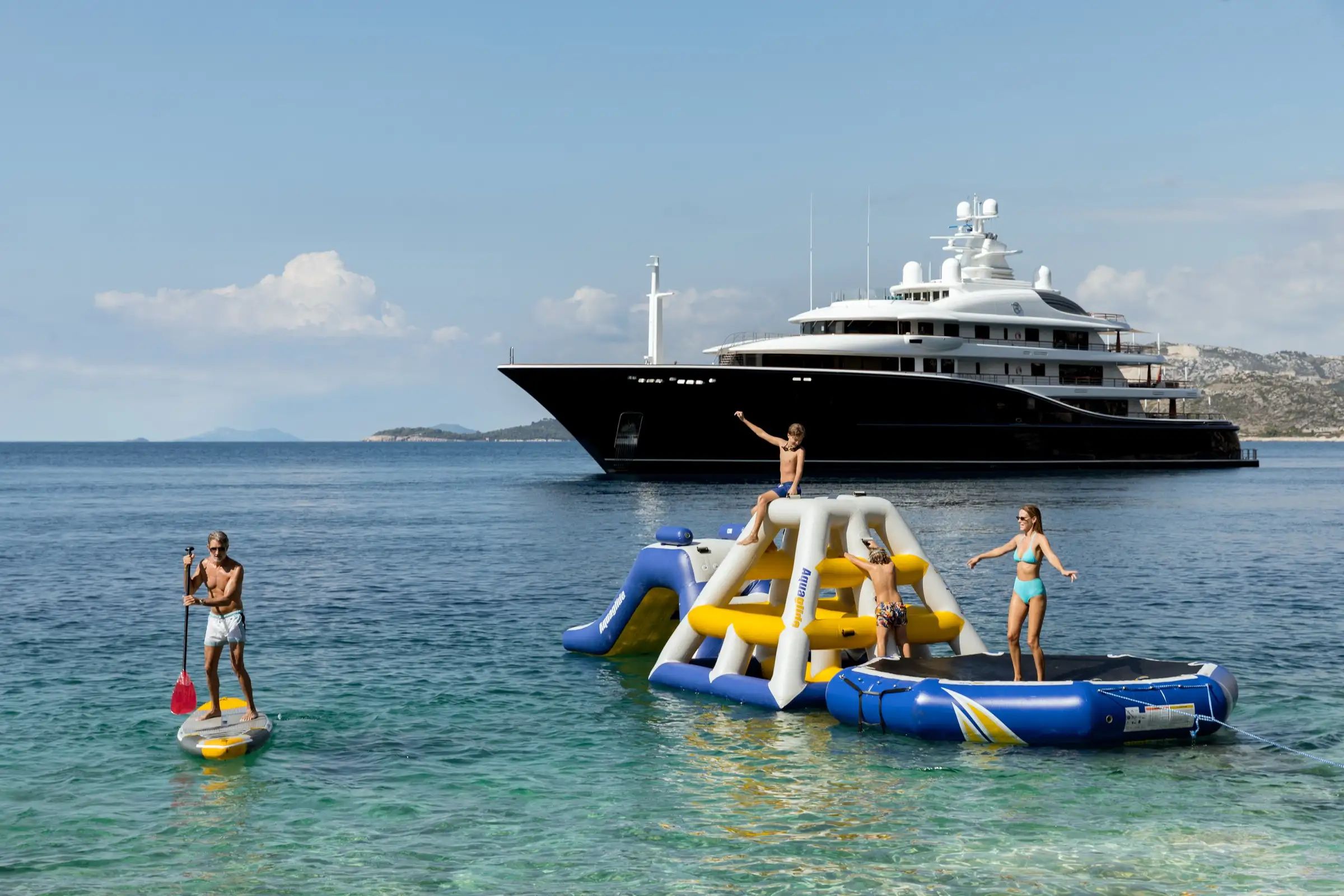 A group of people having fun on inflatable rafts in front of a grand, luxurious Superyacht Aquila on a bright day.