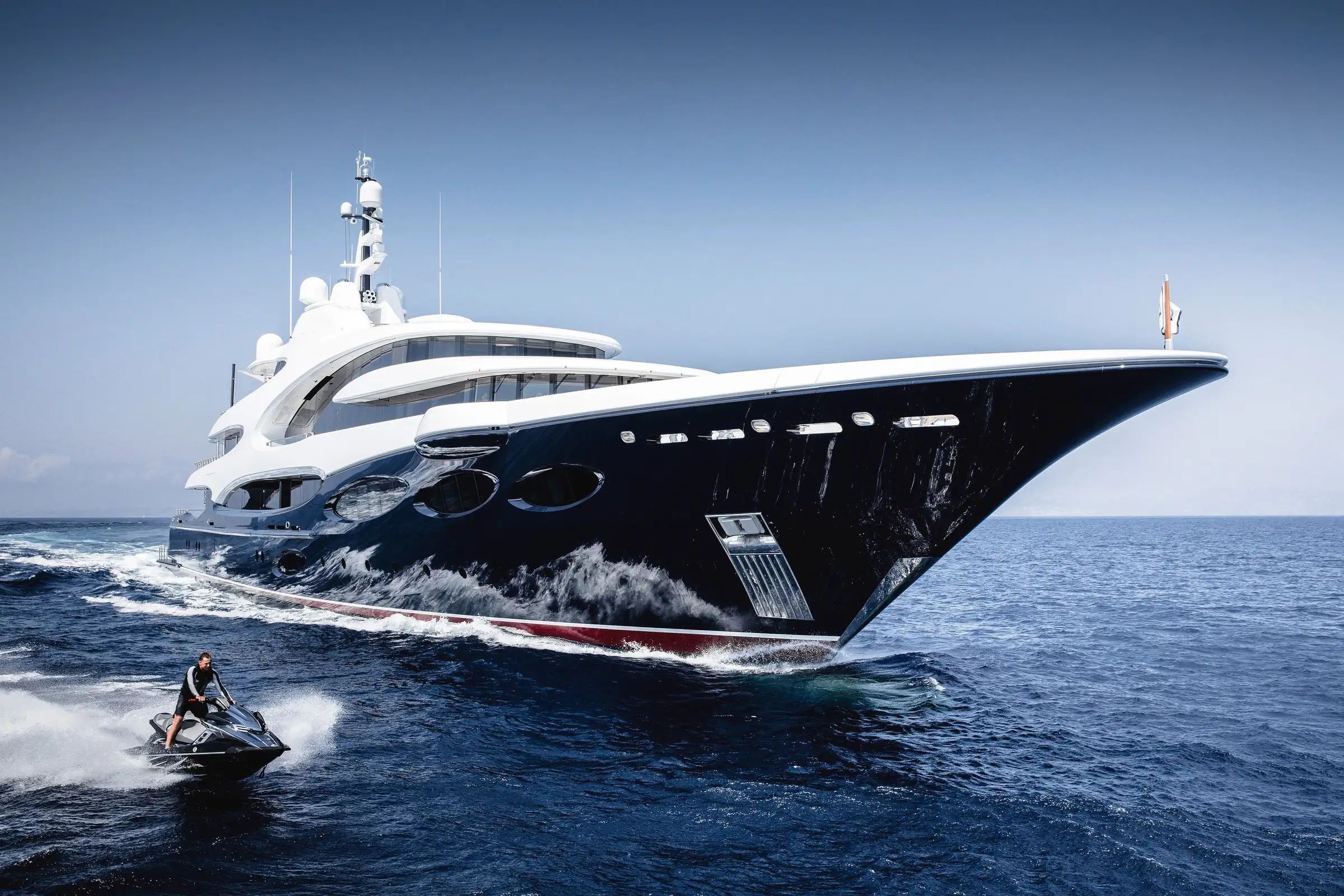 A massive superyacht Custom Line Yacht, BARBARA, sailing stylishly on blue water alongside a jet ski.