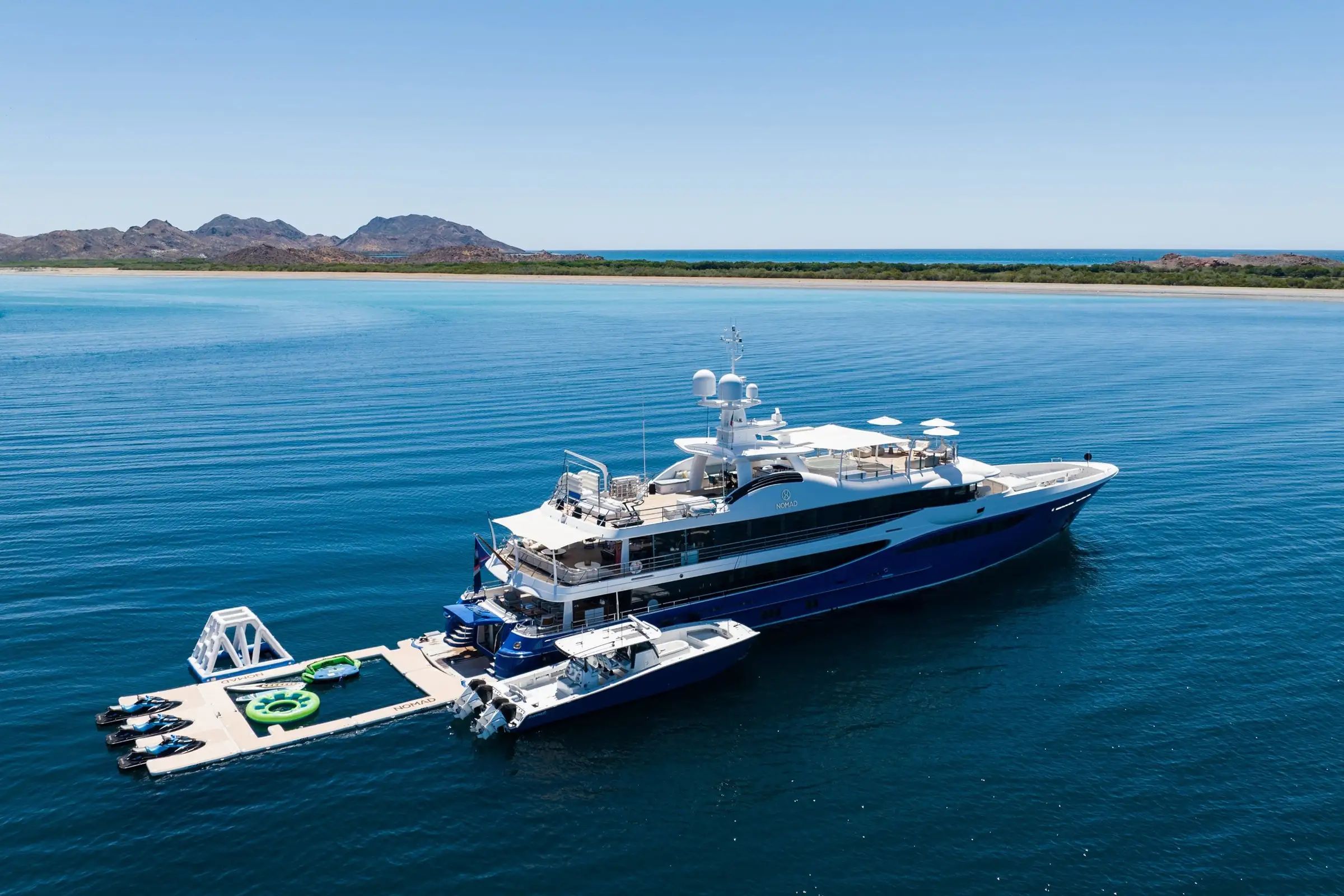 A fancy superyacht Nomad designed in white and blue shown attached to its own seapool and ski boat on the serene waters.