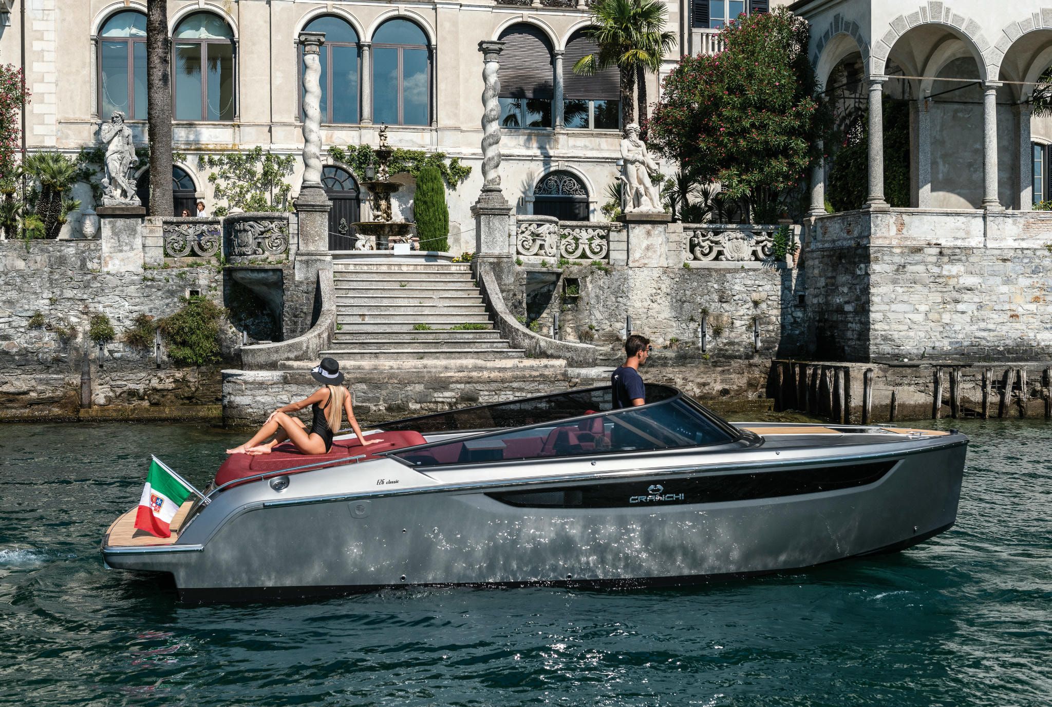 Cranchi Yacht: Grey and red E26 Classic moored in front of an elegant mansion on serene waters.