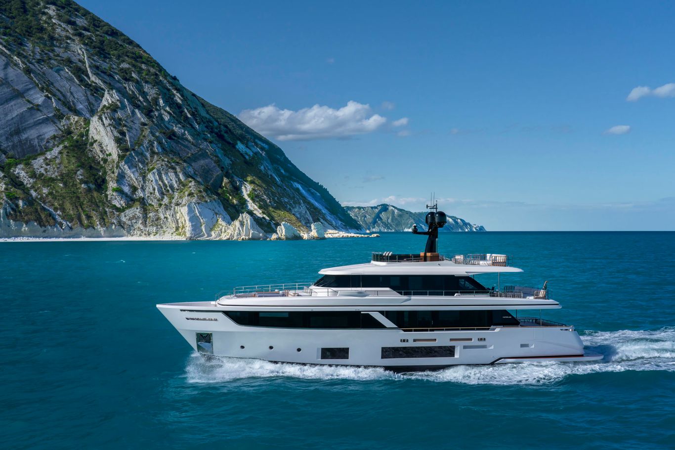 Custom Line Navetta 30 sailing near a rocky cliff through the deep blue ocean.