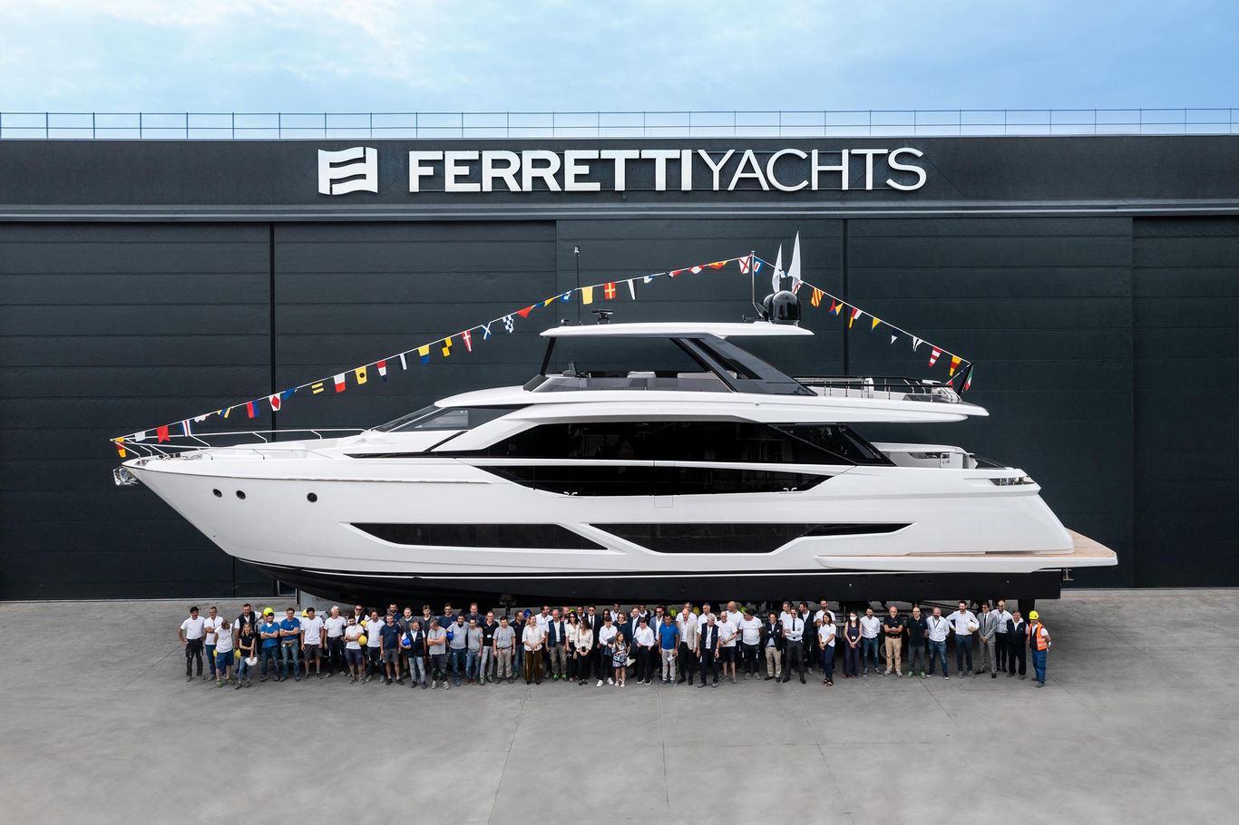 Team photo in front of a Ferretti luxury motor yacht at the shipyard facility, decorated with international flags, by Starship Yachts.