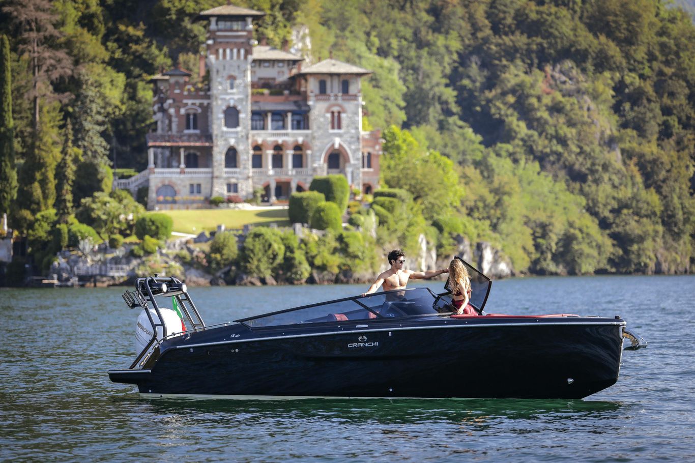 Couple cruising in Cranchi E26 Rider yacht on a calm lake, framed by lush greenery and a grand mansion, by Starship Yachts.
