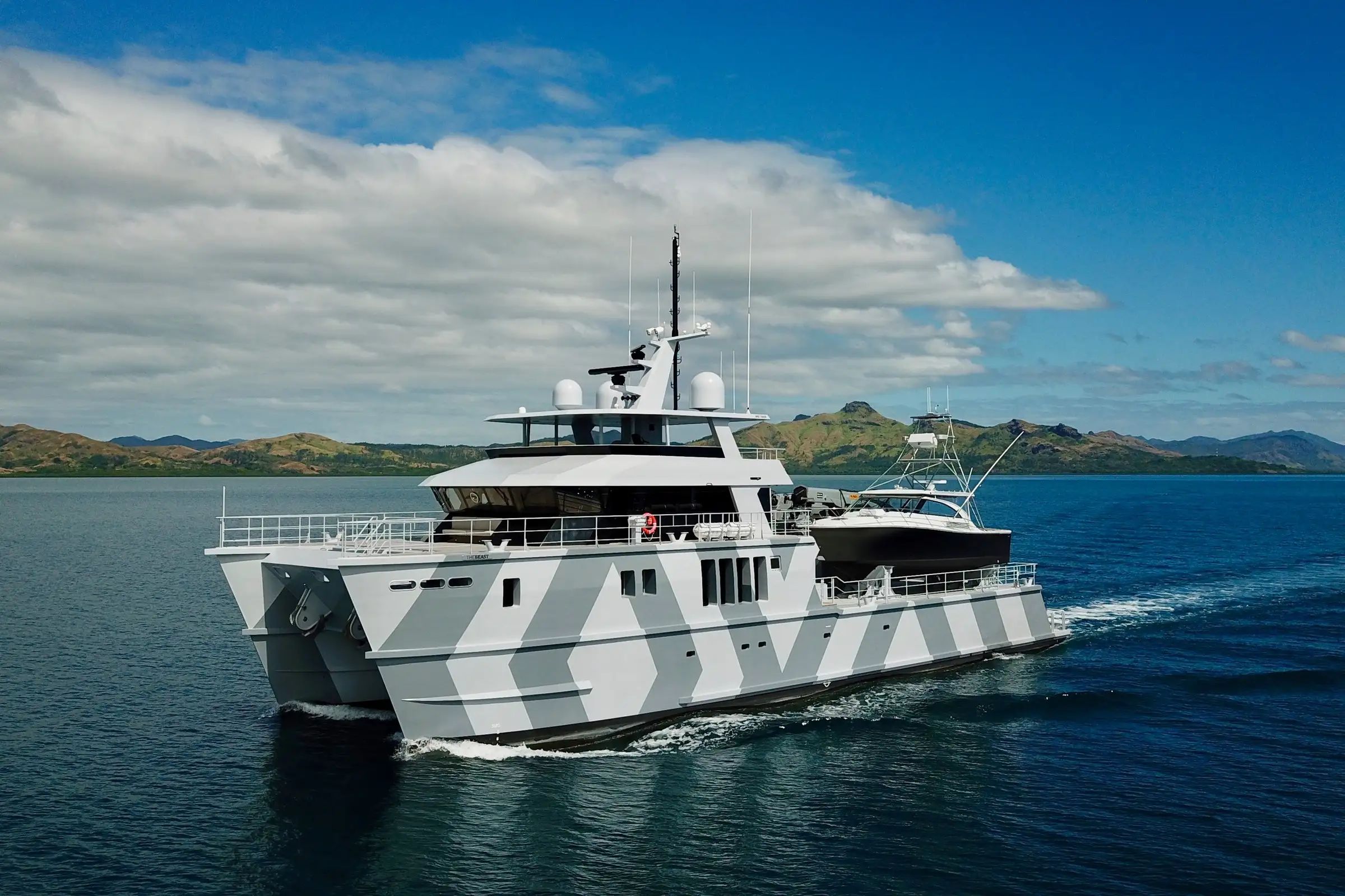 A white, largely built superyacht, The Beast sailing smoothly on serene waters with a mountain view on a bright day.