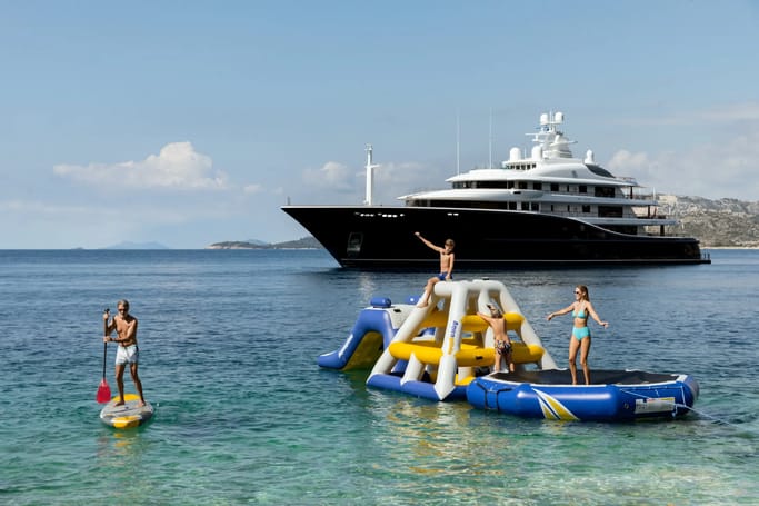 A group of people having fun on inflatable rafts in front of a grand, luxurious Superyacht Aquila on a bright day.