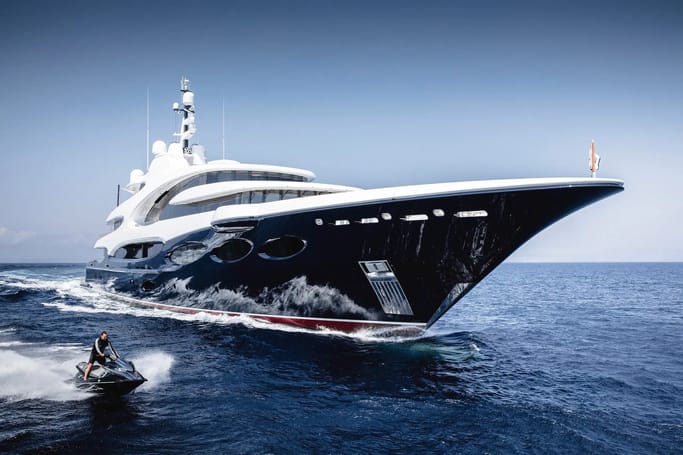 A massive superyacht Custom Line Yacht, BARBARA, sailing stylishly on blue water alongside a jet ski.