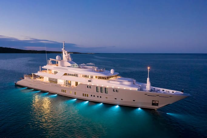 Superyacht Siren with a white hull, lit by turquoise lights, docked at dusk on a calm ocean, with sunset at the background