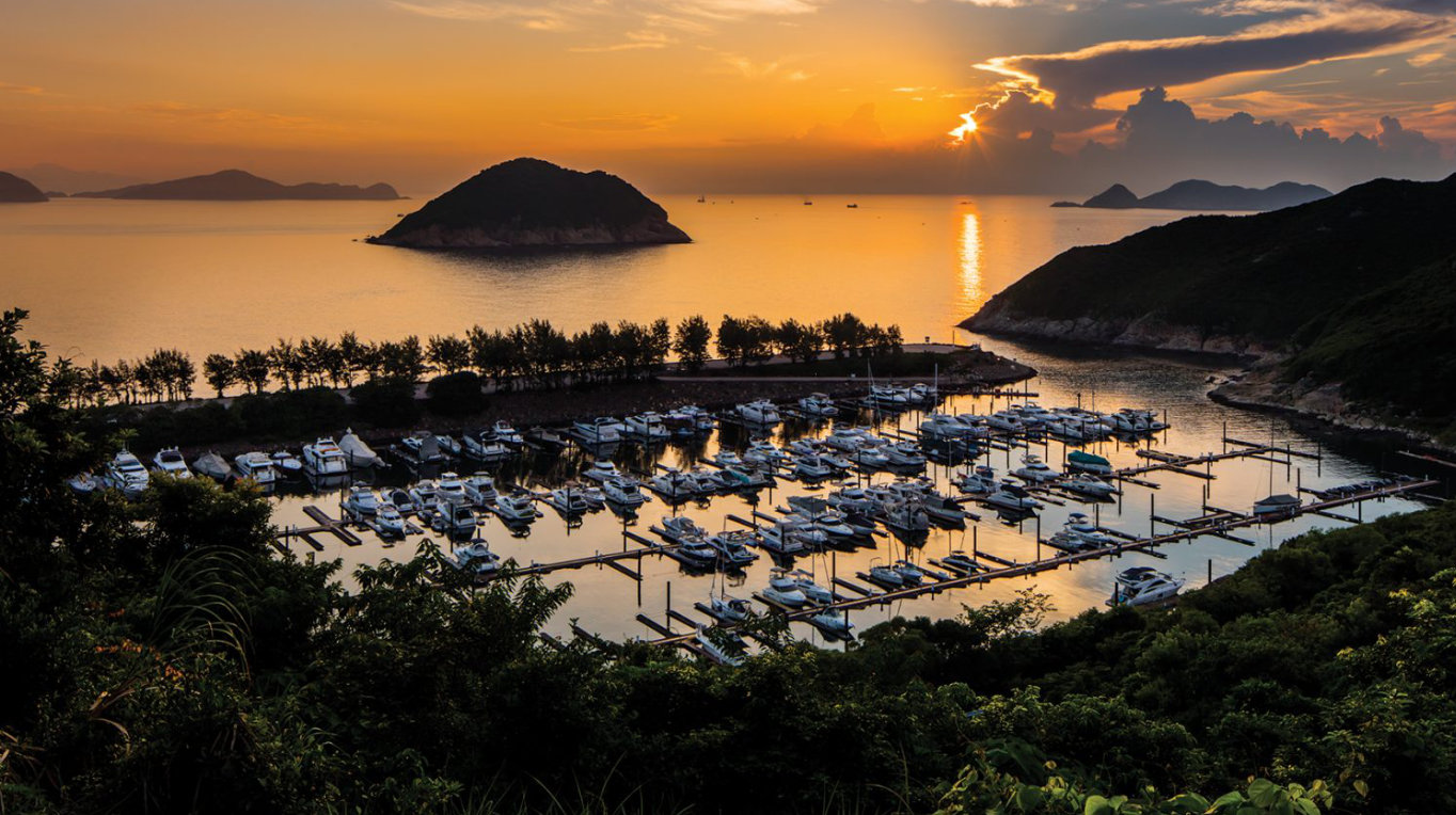 Sunset over Clear Water Bay Marina Club with yachts, islands on horizon, vibrant sky reflections on water, and lush greenery.